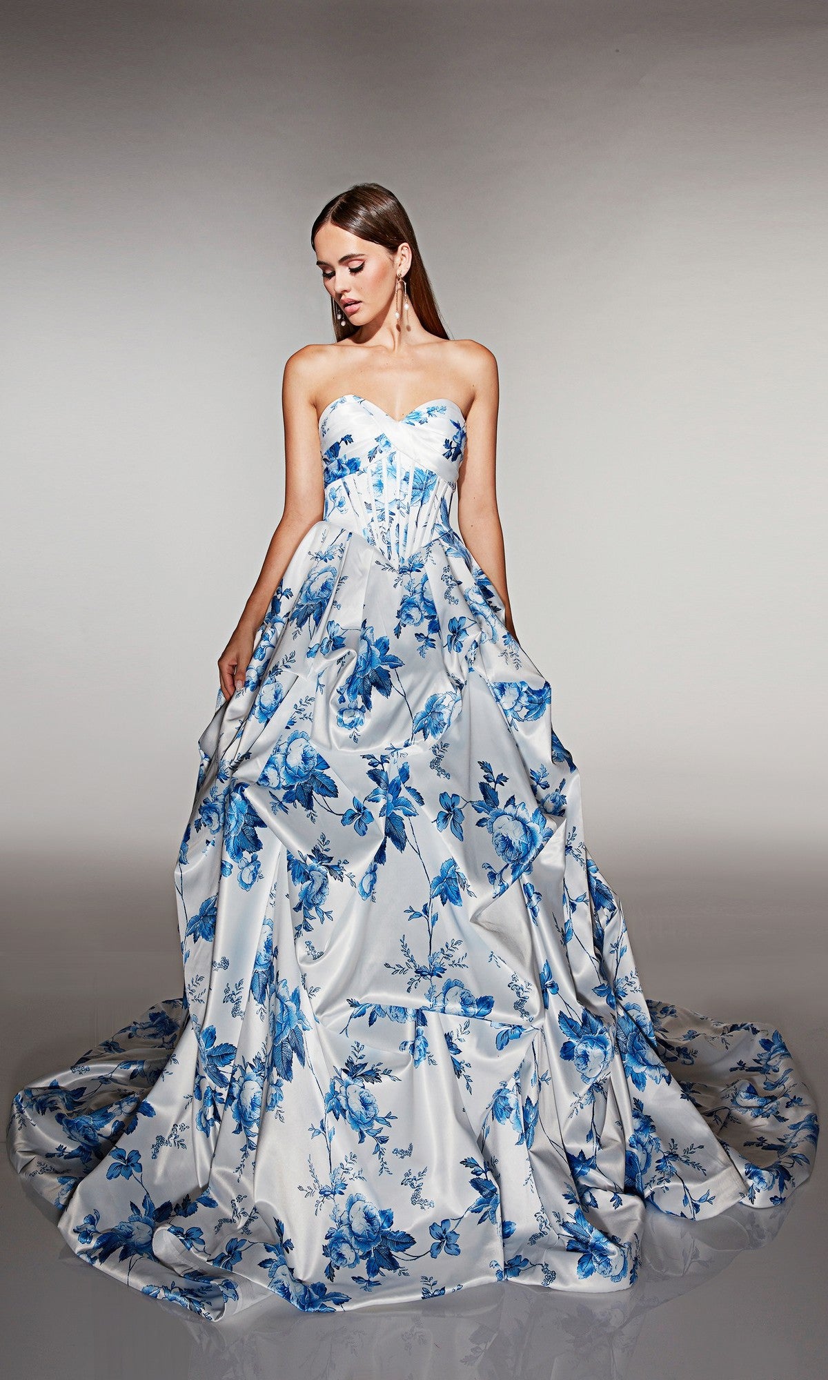 A woman models the Alyce 62213 long prom dressโstrapless, floor-length with a blue floral pattern on a white base and an elegant corset bodiceโstanding against a plain background.